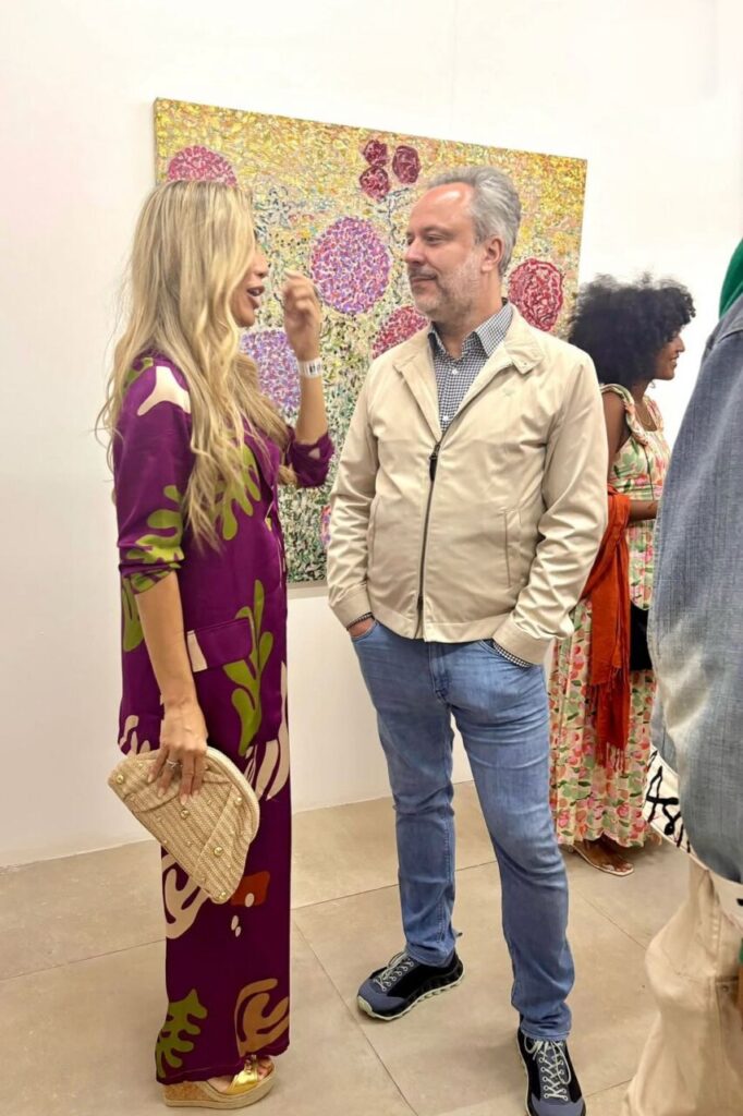 Dos personas conversando en una feria de arte con obras coloridas de fondo.