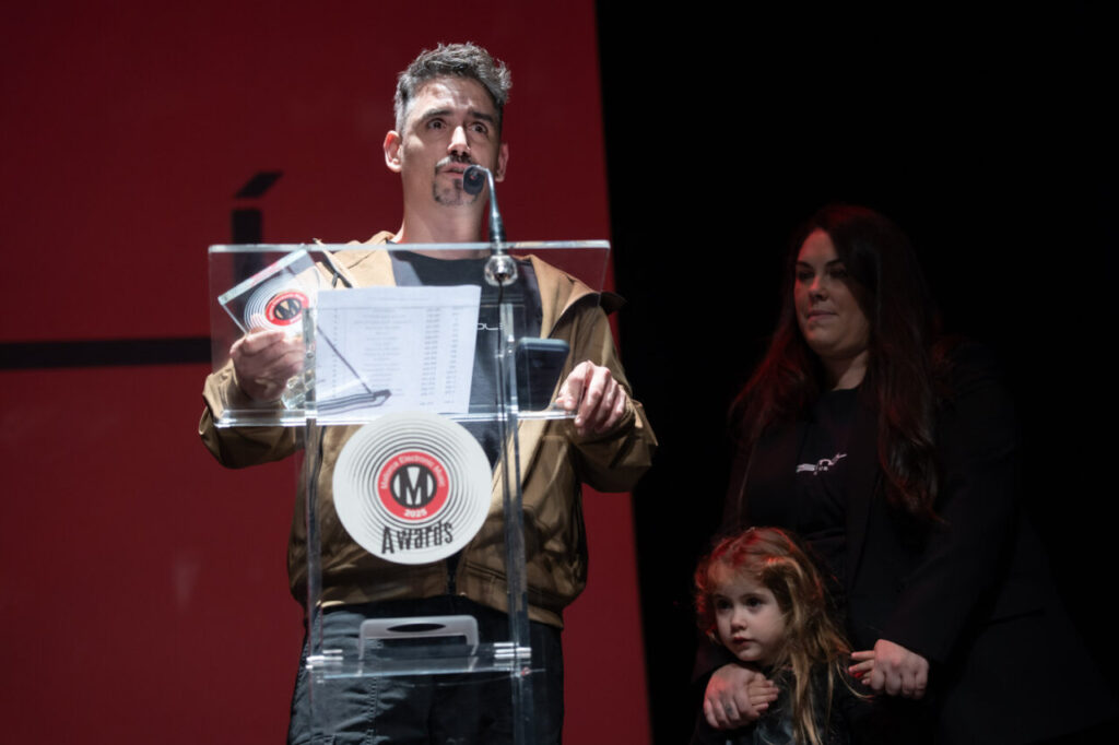 Persona dando un discurso en la Gala MEM Awards con una niña y otra persona al fondo.