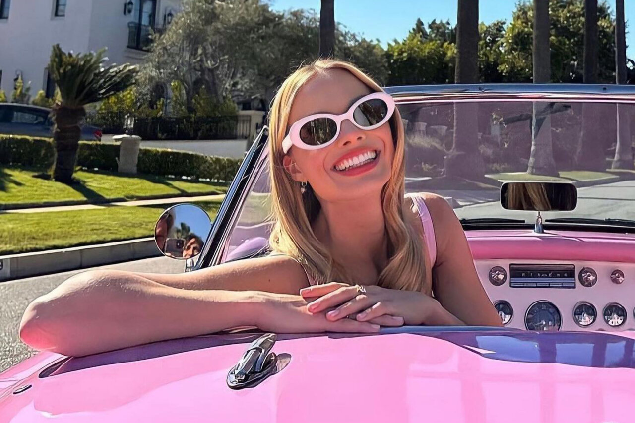 Margot Robbie sonriendo desde un auto rosa con gafas de sol