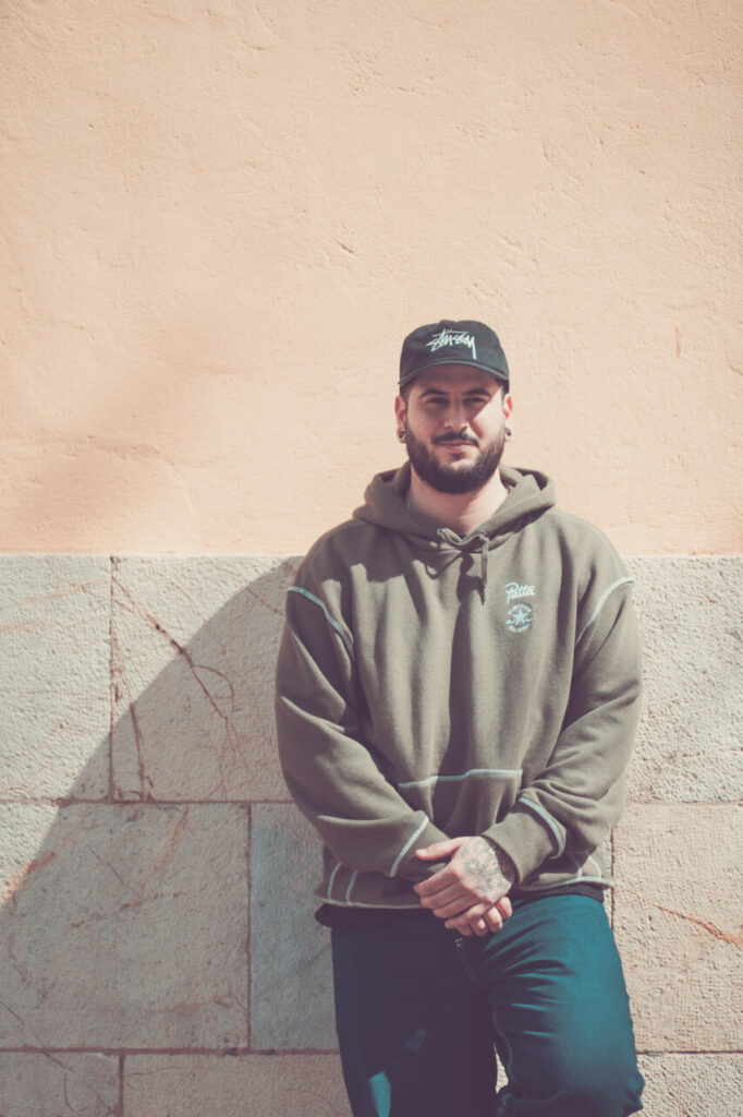 Hombre con sudadera y gorra posando frente a pared de color claro