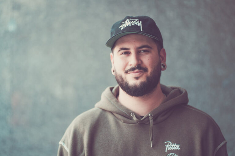 Retrato de un hombre con barba y gorra en un fondo gris.
