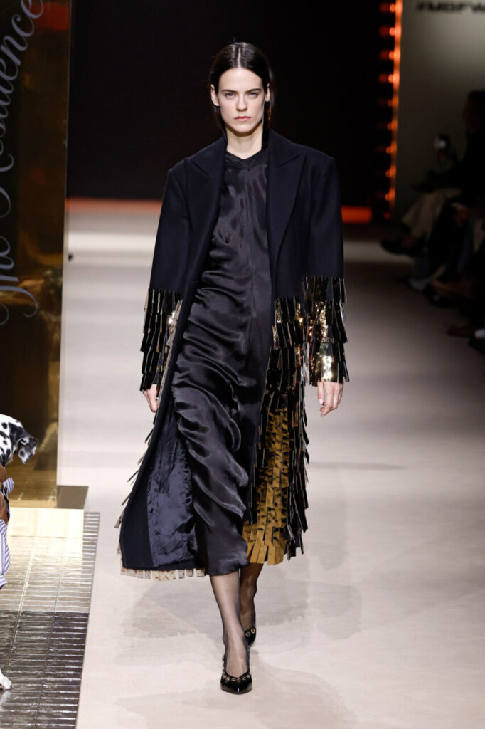 Modelo desfilando con un abrigo negro y vestido brillante en la pasarela.