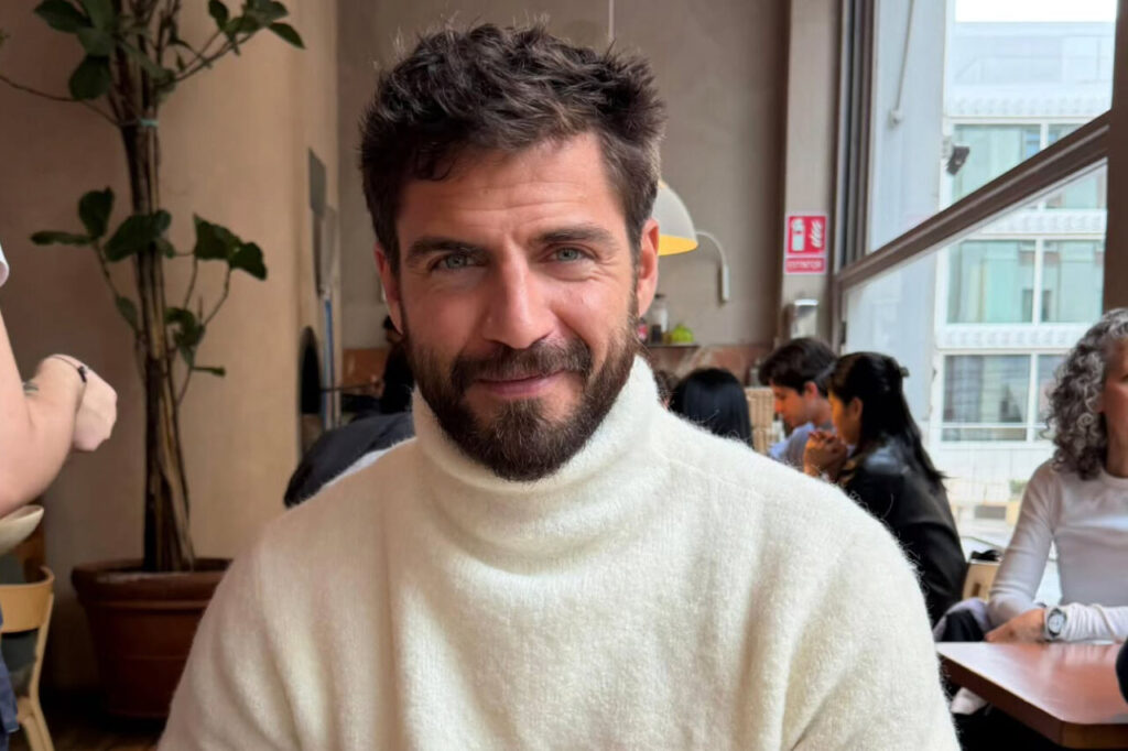 Hombre con barba y suéter blanco sonriendo en una cafetería.