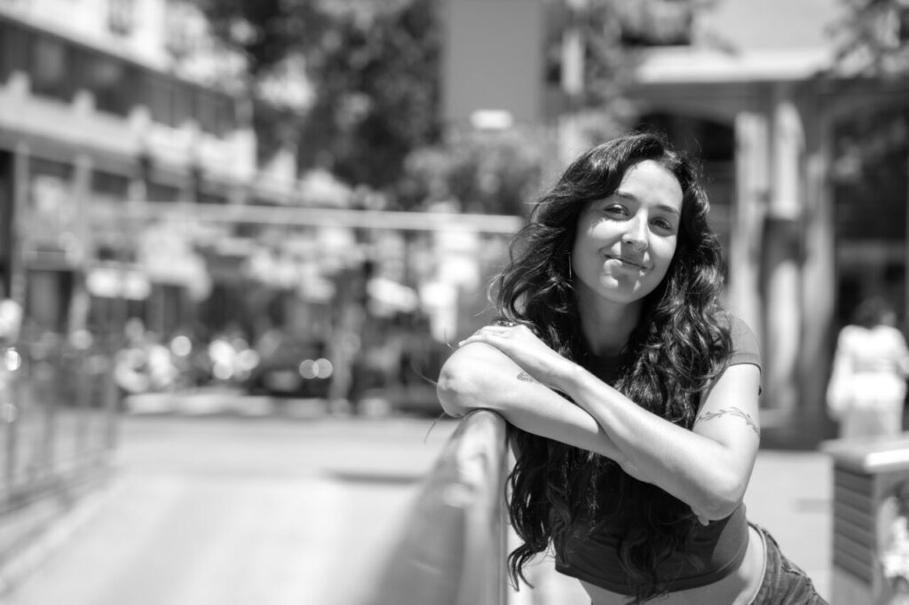 Mujer sonriendo en un entorno urbano, con cabello rizado y brazos cruzados.
