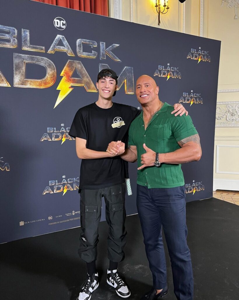 Dos personas posando en un evento de Black Adam, sonriendo y estrechándose la mano.
